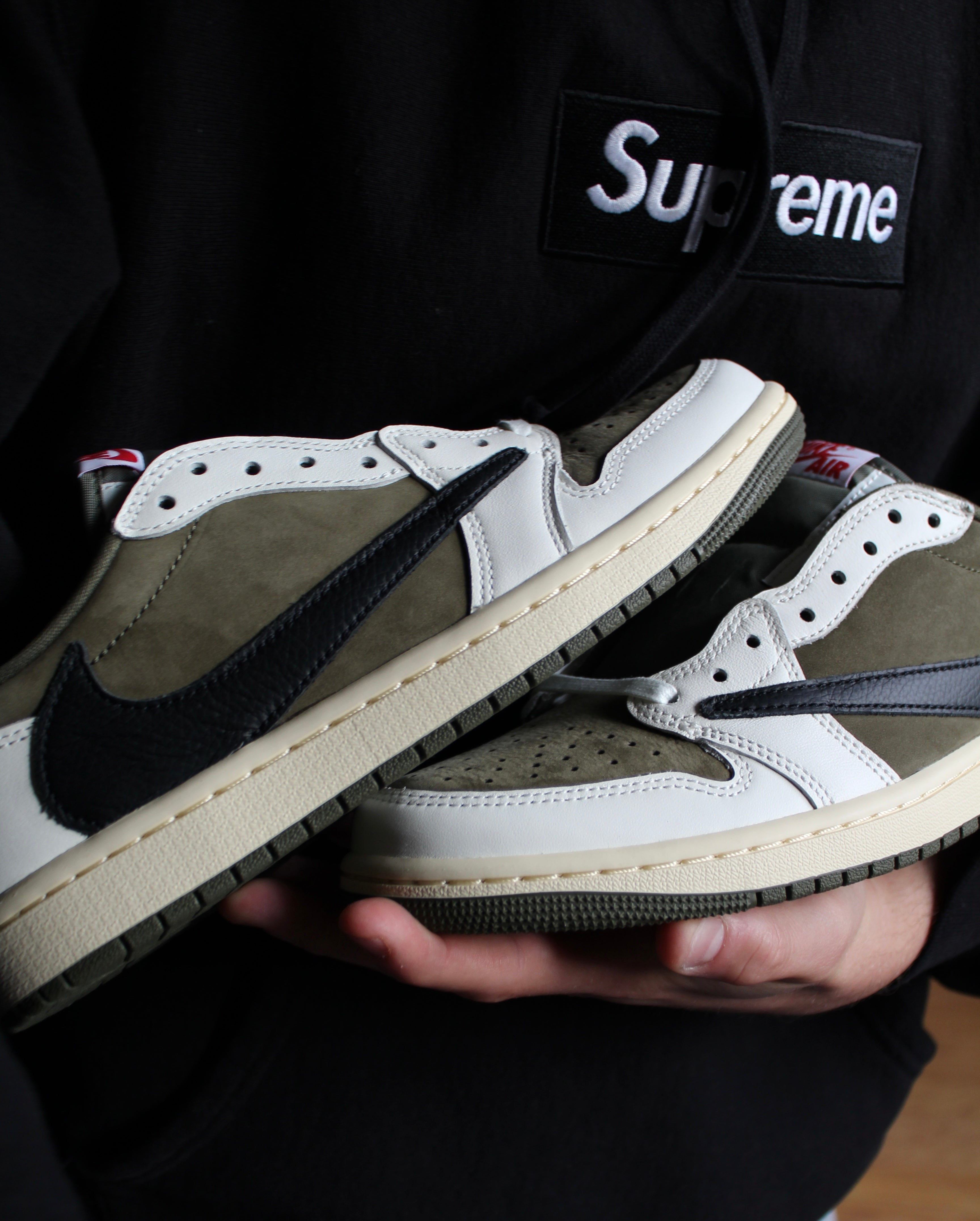 Person holding a pair of Nike sneakers - travis scott, with a visible Supreme logo on a black background